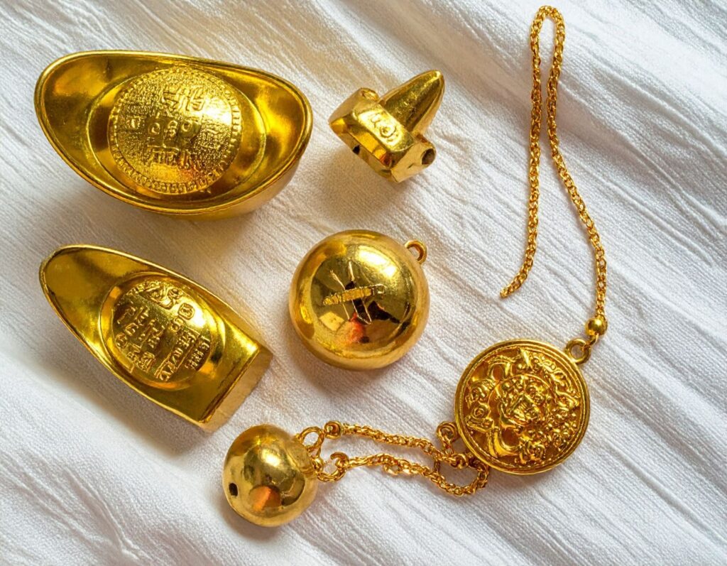 三款彌月金飾平放於絨布背景：金元寶、金鈴鐺與長命鎖造型各具特色